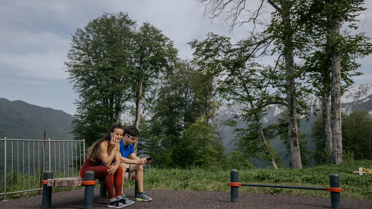 A couple in sports clothing sitting on a bench outdoors, surrounded by trees and mountains.